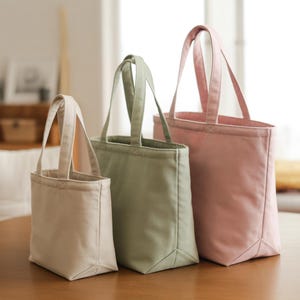 May include: Three canvas tote bags in varying sizes and colors. The smallest is a light beige, the middle one is a muted green, and the largest is a soft pink. Each bag has matching handles and a rectangular shape.