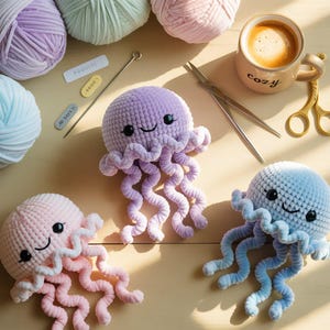 Pode incluir: Medusas de crochê feitas à mão em tons pastel de rosa, lilás e azul, cada uma com um rosto sorridente e tentáculos encaracolados. A imagem inclui novelos de lã, uma chávena de café com a palavra "cozy", tesouras e outros materiais de artesanato.