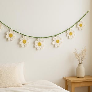 May include: A daisy garland with white petals and yellow centers hangs against a white wall. The garland is strung on a green cord. Below, a bed is made with white linens and pillows. A small wooden table holds a vase with dried flowers.