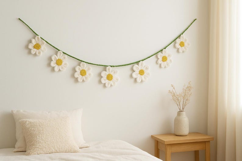 May include: A daisy garland with white petals and yellow centers hangs against a white wall. The garland is strung on a green cord. Below, a bed is made with white linens and pillows. A small wooden table holds a vase with dried flowers.