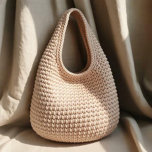 May include: A beige, crocheted handbag with a rounded base and a single, large handle. The bag is made of thick yarn, creating a textured surface. The neutral colour and simple design make it a versatile accessory.