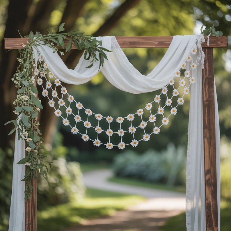 May include: Wooden wedding arch decorated with white fabric, a daisy chain, and greenery. The daisy chain is strung across the arch, and the fabric drapes down. The arch is set up outdoors with a blurred background of trees and a path.