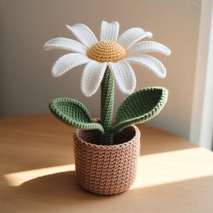 May include: A crocheted daisy flower in a woven pot. The flower has white petals, a yellow center, and a green stem and leaves. The pot is a light brown color, and the entire piece is handmade.