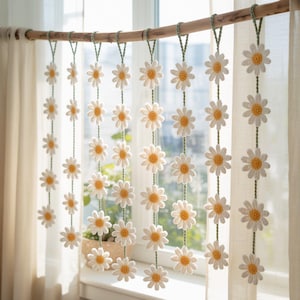 May include: A decorative daisy chain curtain. The curtain features multiple strands of crocheted daisies with white petals and yellow centers. The daisies are suspended from a wooden rod with green yarn. The curtain hangs in front of a window with natural light.