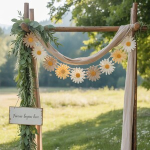 May include: A wooden wedding arch decorated with a daisy garland, greenery, and a draped fabric. A small sign reads "Forever begins here". The daisy garland features white, yellow, and tan crocheted flowers, creating a rustic and romantic aesthetic.
