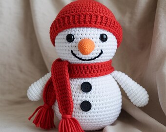 May include: A handmade crochet snowman with a red hat, scarf, and carrot nose. The snowman has black button eyes and a stitched smile. The scarf has tassels. The snowman is a winter holiday decoration.