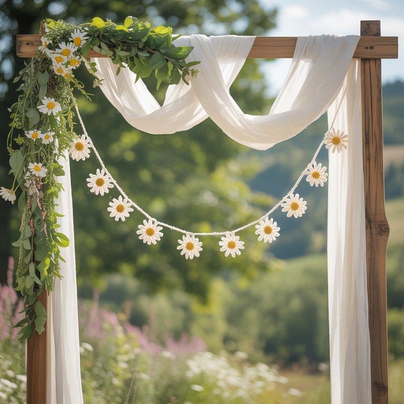 May include: A wooden wedding arch decorated with white fabric, fresh daisies, and greenery. A daisy garland hangs across the front. The background is a blurred view of a green field and trees.