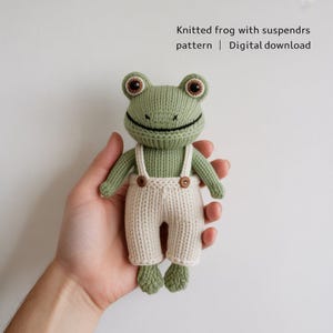 May include: A knitted green frog toy wearing cream-coloured dungarees with wooden button details. The frog has large brown eyes and a smiling black mouth. The text "Knitted frog with suspenders pattern | Digital download" is visible.