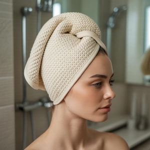 Pode incluir: Uma toalha de cabelo creme, texturizada, enrolada na cabeça de uma pessoa. A toalha é feita de um material espesso e absorvente e tem um laço para prendê-la. A pessoa está olhando para o lado, com uma expressão neutra.