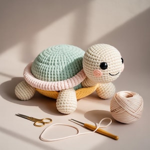 Puede incluir: Una tortuga de ganchillo hecha a mano en colores pastel. La tortuga tiene un caparazón azul claro, un borde rosa y amarillo, y cabeza y extremidades color crema. Ojos negros y boca sonriente. También hay un ovillo de lana, tijeras y un ganchillo.
