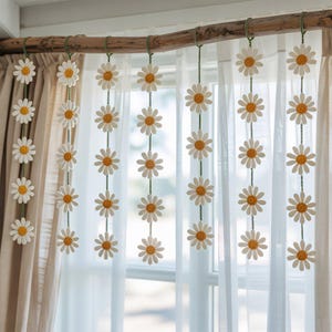 May include: Daisy flower garland hanging from a wooden rod. The garland features white petals with yellow centers, strung on green cords. The daisies are displayed in front of a sheer white curtain.