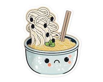 May include: A blue bowl with a cute cartoon face holds a serving of ramen noodles with black sesame seeds and a green garnish. A pair of chopsticks sticks out of the bowl.