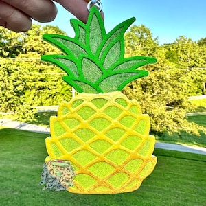 May include: A yellow and green pineapple-shaped charm with glitter accents. The charm is suspended from a beaded yellow string. The charm has a green top and a yellow body. The charm has the text "Burgo Creek Creations".