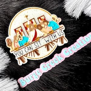 May include: A white round ceramic ornament with a depiction of the Last Supper and the text "You Can Sit With Us". The ornament is decorated with gold trim and has a pink and blue background.