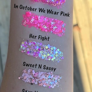 May include: Five different glitter samples on a person's arm. The samples are labeled "In October We Wear Pink", "Her Fight", "Sweet N Sassy", "Save the TaTas", and "Disco Barbie".
