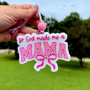 May include: A white felt ornament featuring the pink embroidered text So God made me a MAMA with a pink bow graphic. The ornament is attached to a white elastic cord with a textured pink bead, creating a decorative hanging accessory.