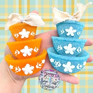 May include: Two stacks of three nested bowls in bright orange and light blue, each featuring a textured surface and a white floral relief design. Each stack is topped with a frayed beige fabric bow. A logo with the text Burge Creek Creations is visible.