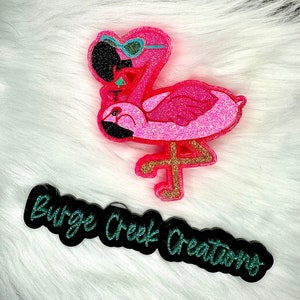 May include: A bright pink glittery flamingo-shaped ornament wearing teal-rimmed sunglasses. Below the flamingo is a black plaque with the text Burge Creek Creations written in a teal glittery script font, featuring a distinct decorative design.