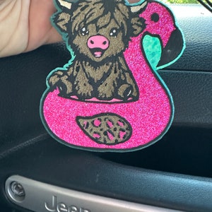 May include: A pink glitter flamingo pool float with a brown and black Highland cow sitting inside. The float has a leopard print design on the bottom.