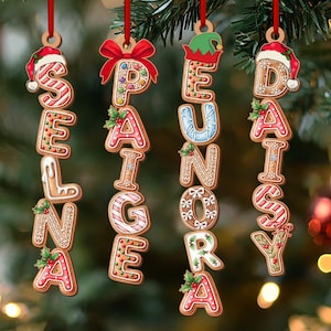 May include: Gingerbread-style Christmas ornaments spelling out "SEASON'S DAY". Each letter is decorated with icing, holly, and festive hats. The ornaments hang from red ribbons against a blurred background of Christmas tree branches and lights.