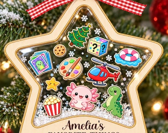 May include: A star-shaped wooden Christmas ornament with a clear center filled with small white beads. Inside are colorful illustrations of a tree, question block, life ring, helicopter, axolotl, popcorn, palette, and cards. Text reads: Amelia's FAVORITE THINGS 2026.