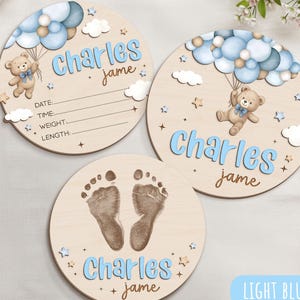 May include: Three round wooden baby milestone plaques. Two plaques feature the name "Charles" in light blue, with a teddy bear and balloons. The third plaque has baby footprints and the name "Charles". The text "jame" is also present.