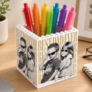 May include: A white pen holder with a "Best Dad Ever" design and photos of two children. The holder is filled with a rainbow of pens. The pen holder is on a wooden desk.