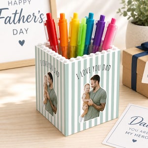 May include: A personalized pen holder with a photo and the text "I LOVE YOU DAD" and "Dad YOU ARE MY HERO". The holder contains a rainbow of pens. A framed sign says "HAPPY Father's DAY". A gift box and card are also present.