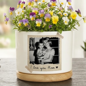 May include: A white ceramic flower pot with a black and white photo of a mother and child, and the text "I love you Mom". The pot is filled with colorful pansies and daisies. The pot sits on a wooden base.