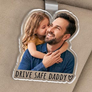 May include: A clear acrylic car visor charm featuring a photo of a man and a child. The child is kissing the man's cheek. The text "DRIVE SAFE DADDY" is printed below the photo. The charm is attached to the visor with a metal clip.