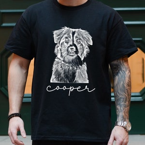 May include: Black t-shirt featuring a detailed black and white illustration of a dog. The dog's name, "cooper", is written in a cursive font below the image. The shirt is a classic crew neck style.