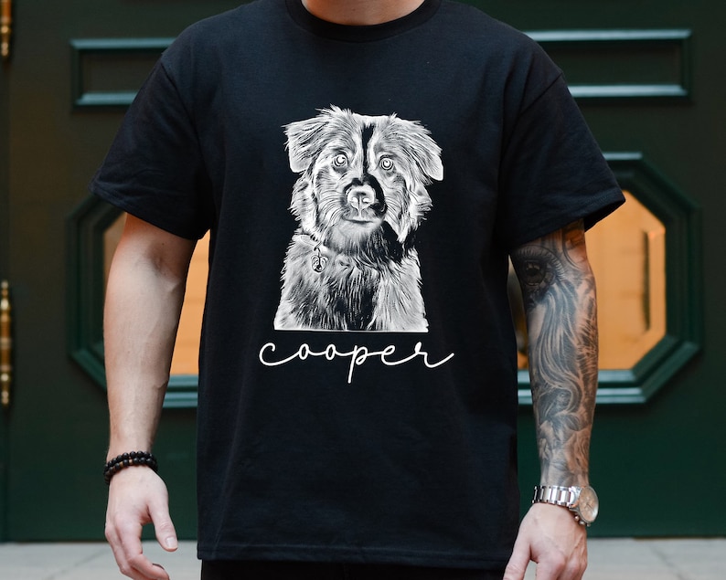 May include: Black t-shirt featuring a detailed black and white illustration of a dog. The dog's name, "cooper", is written in a cursive font below the image. The shirt is a classic crew neck style.