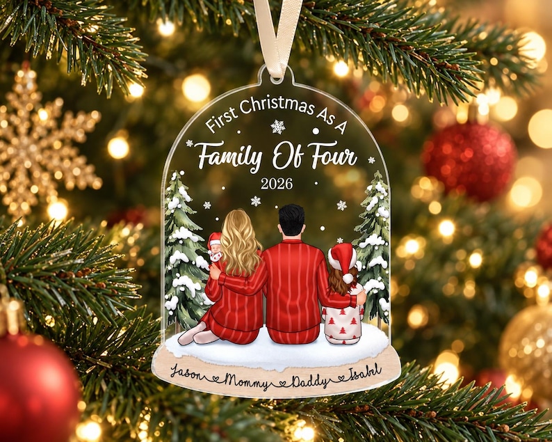 May include: A clear, bell-shaped acrylic Christmas ornament featuring an illustration of a family of four sitting in front of pine trees. The text reads First Christmas As A Family Of Four 2026, with the names Jason, Mommy, Daddy, and Isabel below.