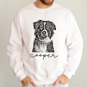 May include: White sweatshirt featuring a black and white illustration of a dog's face. The dog has a collar and the name "cooper" is written in a script font below the image. The sweatshirt is a casual style.