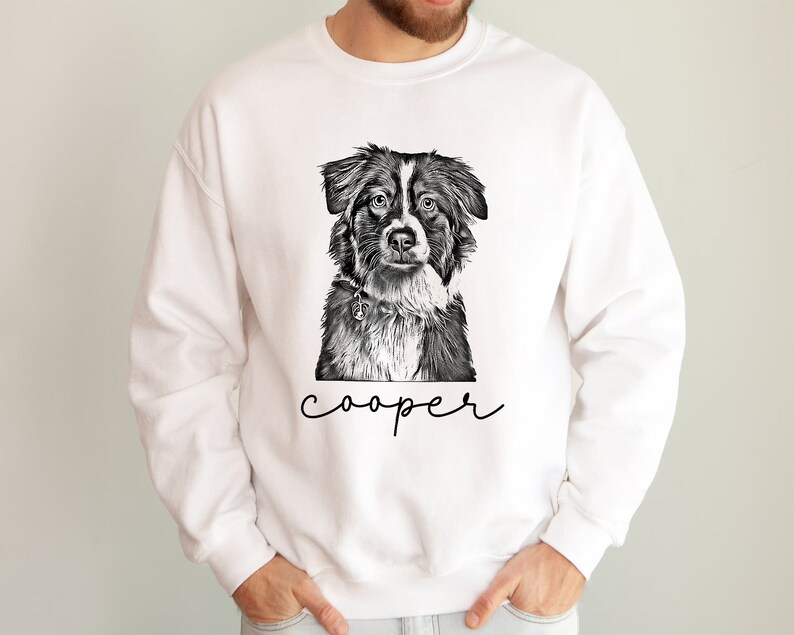 May include: White sweatshirt featuring a black and white illustration of a dog's face. The dog has a collar and the name "cooper" is written in a script font below the image. The sweatshirt is a casual style.
