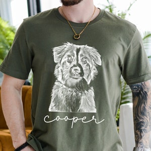 May include: Olive green t-shirt featuring a detailed black and white illustration of a dog's face. The dog has a collar with a charm. The name "cooper" is written in a cursive font below the dog's image. The shirt is made of a soft material.