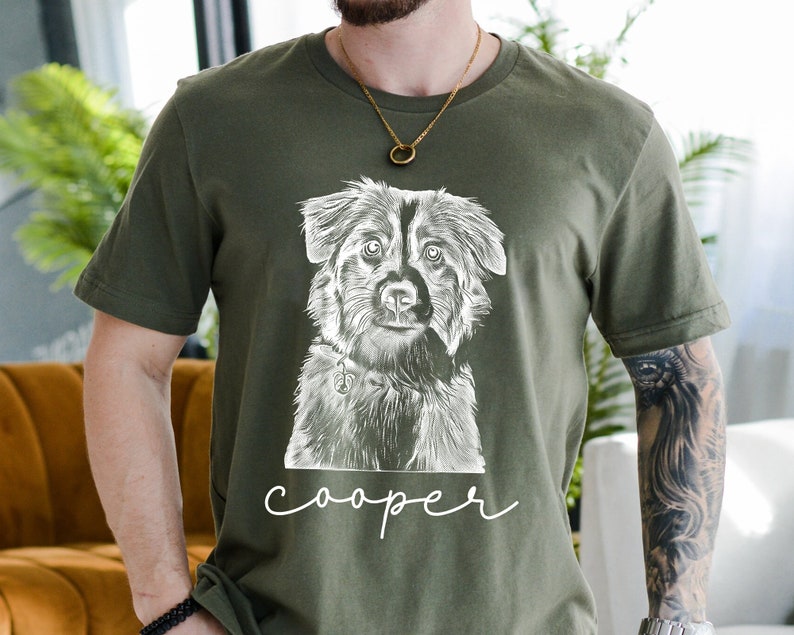 May include: Olive green t-shirt featuring a detailed black and white illustration of a dog's face. The dog has a collar with a charm. The name "cooper" is written in a cursive font below the dog's image. The shirt is made of a soft material.