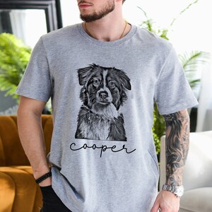 May include: A gray t-shirt featuring a black and white illustration of a dog's face. The dog has a focused expression. The name "cooper" is written in cursive below the dog's image. The shirt is made of a soft material.