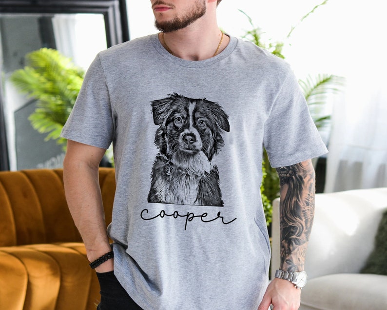 May include: A gray t-shirt featuring a black and white illustration of a dog's face. The dog has a focused expression. The name "cooper" is written in cursive below the dog's image. The shirt is made of a soft material.
