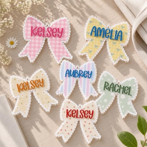 May include: Six decorative bow-shaped ornaments featuring various patterns and colors. The bows are personalized with the names Kelsey, Amelia, Aubrey, and Rachel in raised lettering. Each bow has a scalloped edge and a small loop for hanging.
