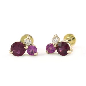 May include: A pair of gold earrings with three gemstones each. The earrings feature a large round ruby, a smaller round pink sapphire, and a small round diamond.