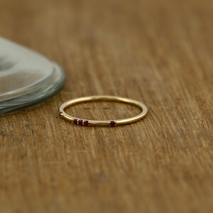 May include: A thin gold band ring featuring four small, round, deep red gemstones set along the top curve. The metal has a polished finish, and the stones are arranged in a cluster of three followed by a single stone spaced slightly apart.