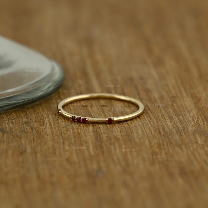 May include: A thin gold band ring featuring four small, round, deep red gemstones set along the top curve. The metal has a polished finish, and the stones are arranged in a cluster of three followed by a single stone spaced slightly apart.