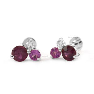 May include: A pair of silver stud earrings with three gemstones each. The earrings feature two round pink sapphires and one round white diamond.
