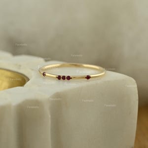 May include: A thin gold band ring featuring four small, round, deep red rubies set along the top. The ring has a polished metallic finish and a simple, slender design. The text Facetssllc is visible in multiple locations across the image.