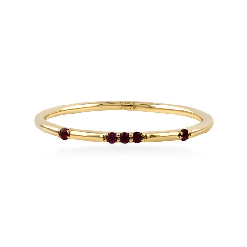 May include: A thin 10K gold band ring featuring five small, round, deep red rubies set along the front. The metal is polished and smooth, with the text 10K clearly engraved on the inner surface of the band, showcasing a simple, delicate design.