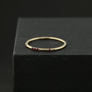 May include: A thin gold band ring featuring four small, round, red gemstones set along one side. The ring has a polished metallic finish and a simple, circular design, showcasing a delicate construction with evenly spaced red stones.