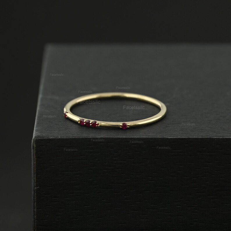 May include: A thin gold band ring featuring four small, round, red gemstones set along one side. The ring has a polished metallic finish and a simple, circular design, showcasing a delicate construction with evenly spaced red stones.