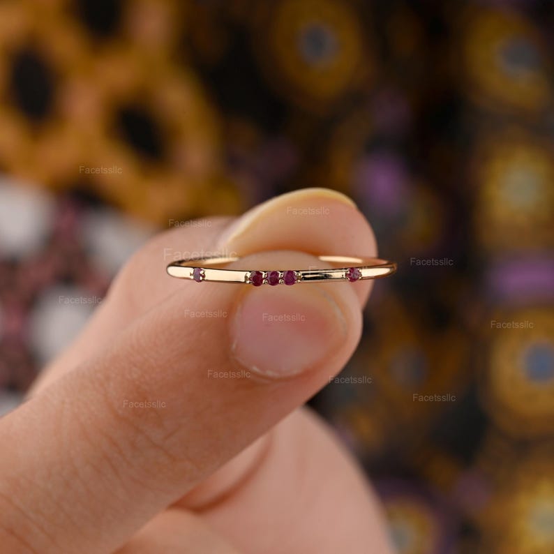 May include: A thin gold band ring featuring five small, round, deep red rubies set horizontally across the front. The ring has a polished metallic finish and a delicate, minimalist construction, showcasing the vibrant red gemstones against the gold.