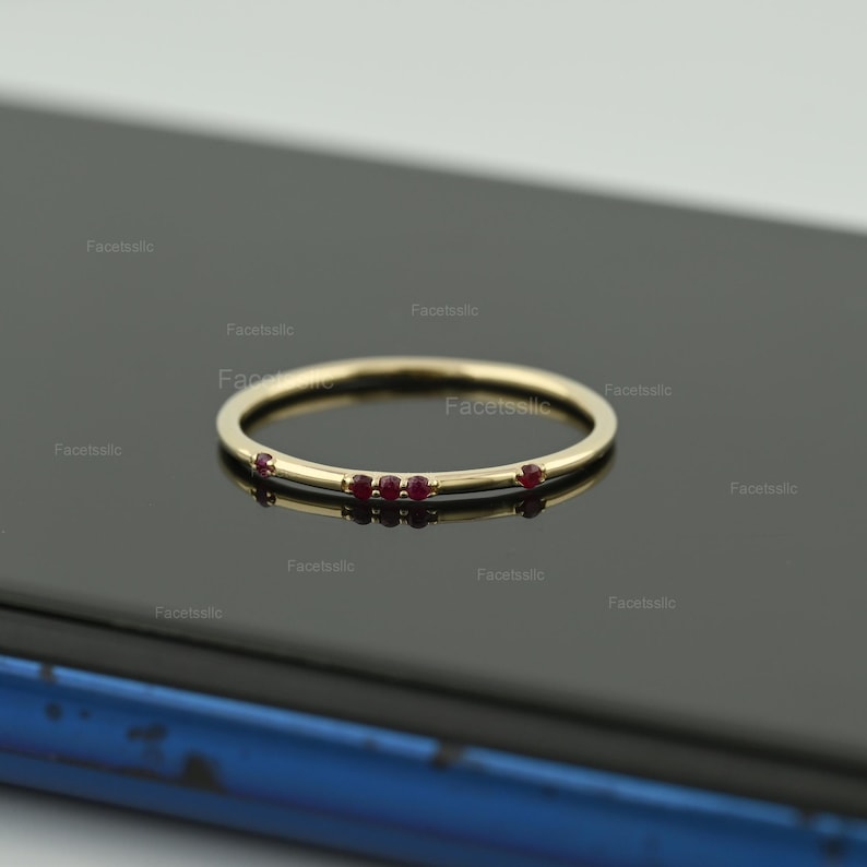 May include: A thin gold band ring featuring four small, round, deep red rubies set along the top curve. The ring has a polished metallic finish and a simple, slender design that highlights the evenly spaced gemstones embedded in the metal band.